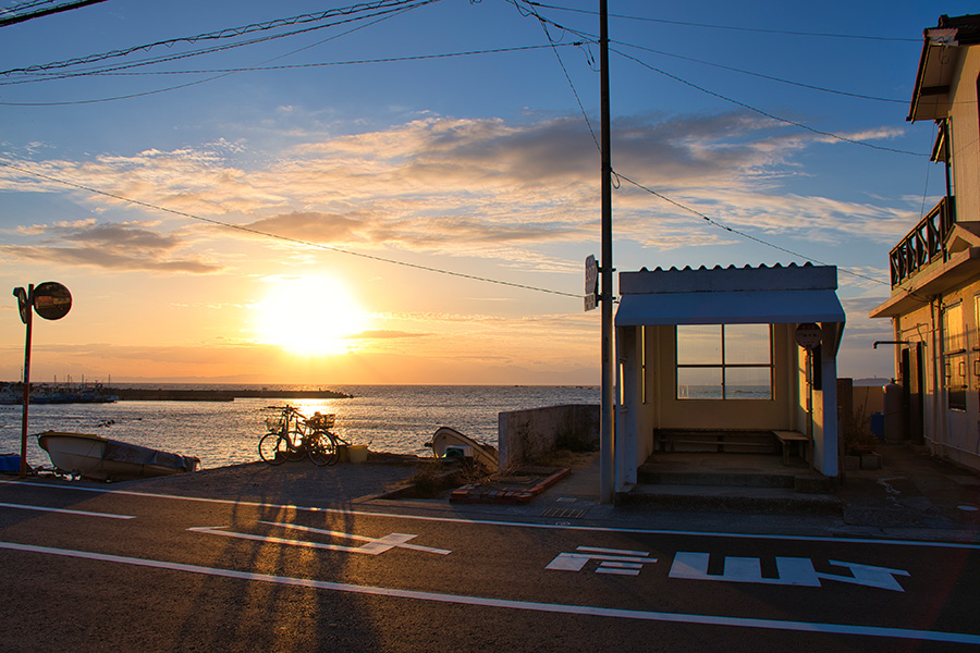 夕暮れの海沿いの町。漁港のそばに佇むバス停と小舟、穏やかな海